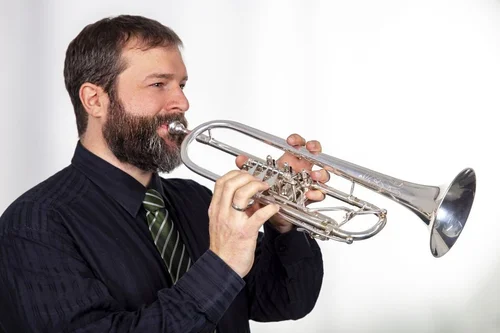 Seitliches Portrait von dem Posaunchorleiter Florian Pöschel im schwarzen Hemd und er hält eine Trompete.